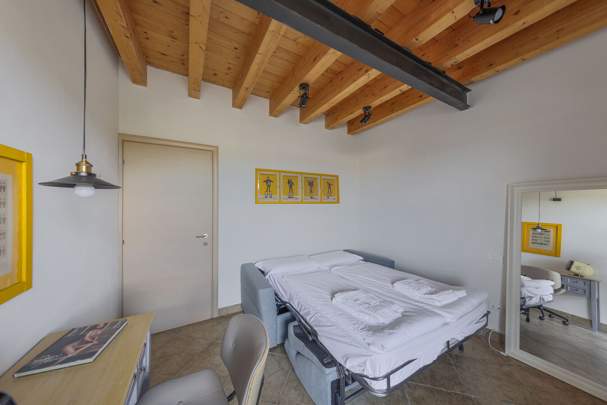 A compact bedroom with a bed, a desk, and a chair. There is a door and a mirror on the wall.