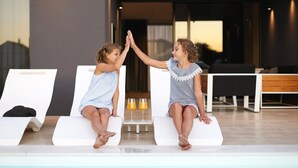 Zwei Personen sitzen auf weißen Liegestühlen am Pool, dazwischen steht ein Tisch mit einem Glas Orangensaft.