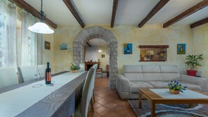 A dining area with a long table, chairs, and a wine bottle. There is a sofa and a coffee table with a plant. An archway leads to another room. - Prisedi in Rakalj With 4 Bedrooms and 3 Bathrooms (Marcana)
