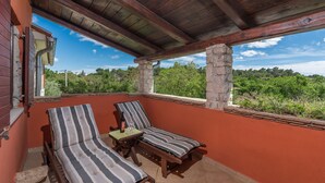 A covered balcony with two striped lounge chairs, a small table with a bottle and glass, and a view of a green landscape. - Prisedi in Rakalj With 4 Bedrooms and 3 Bathrooms (Marcana)