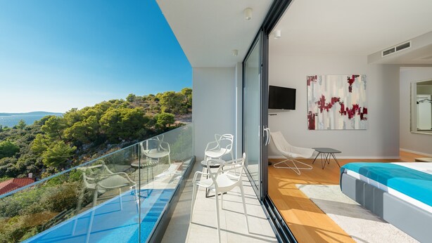 A balcony with a glass barrier, white chairs, a small table, and a bed with a blue cover.