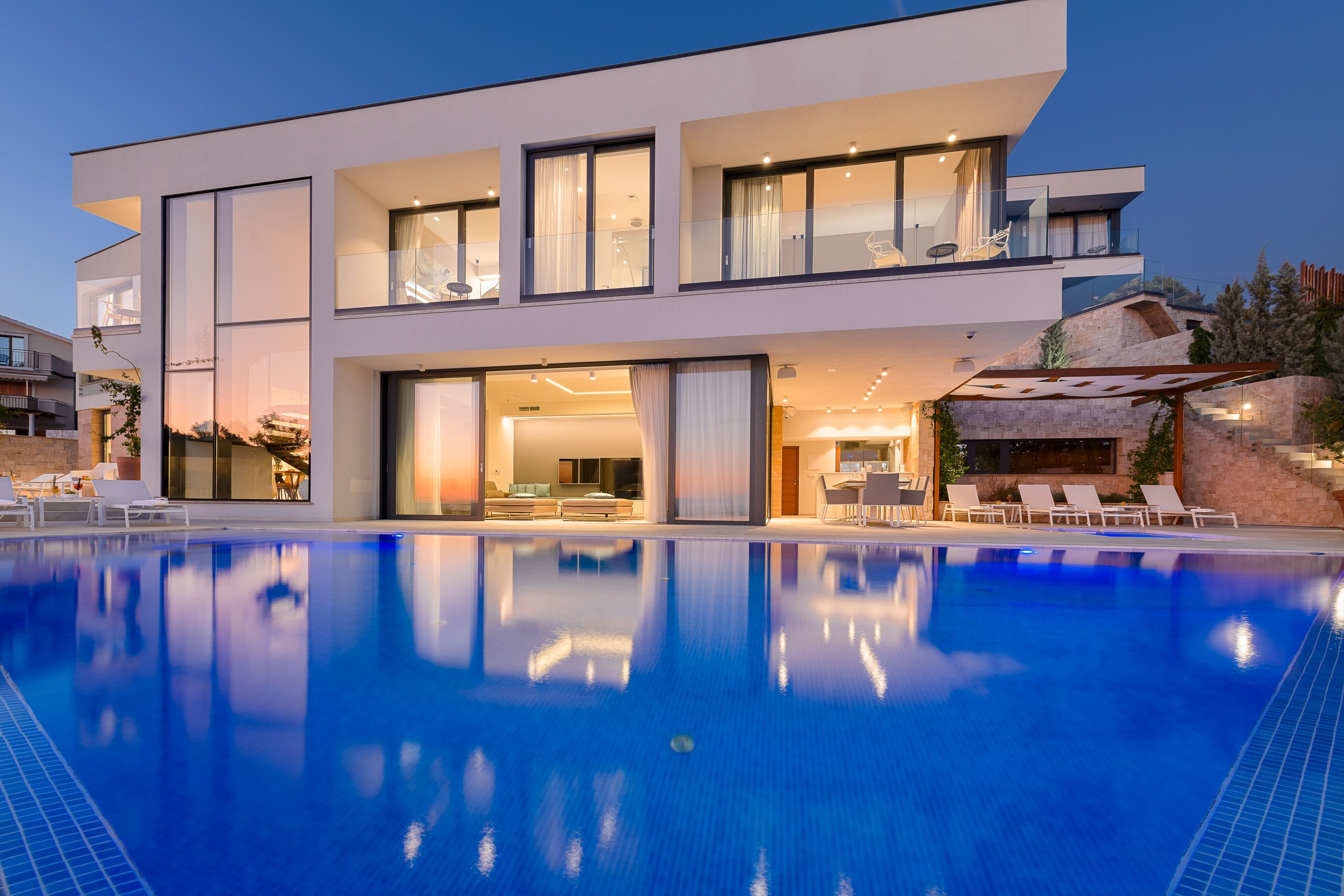 Una casa moderna de dos pisos con piscina, ventanas amplias y mobiliario de exterior.