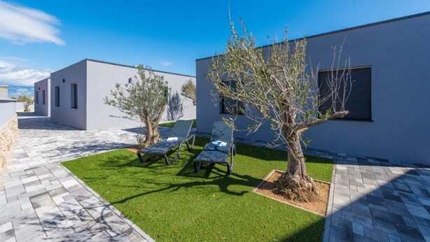 Un espacio exterior moderno con un patio empedrado, un pequeño césped y un árbol. Se observan sillones de descanso y un edificio con tejado plano y amplias ventanas.