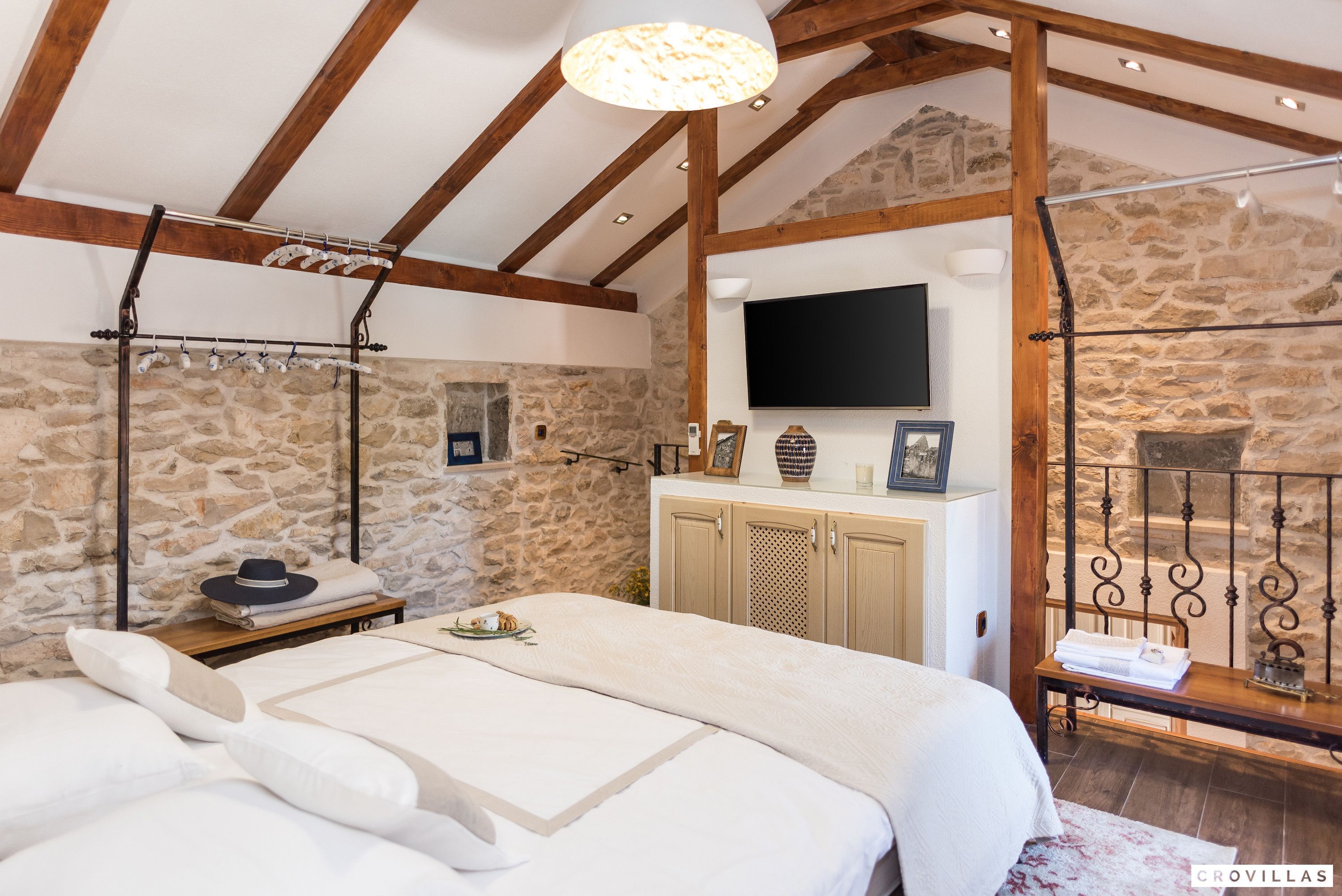 A bedroom with a stone wall, wooden beams, a flat-screen TV, a bed with white linens, a nightstand, and a hanging clothes rack.