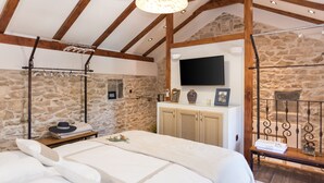 A bedroom with a stone wall, wooden beams, a flat-screen TV, a bed with white linens, a nightstand, and a hanging clothes rack. - Ij Dvori in Skradin With 5 Bedrooms and 5 Bathrooms (Skradin)