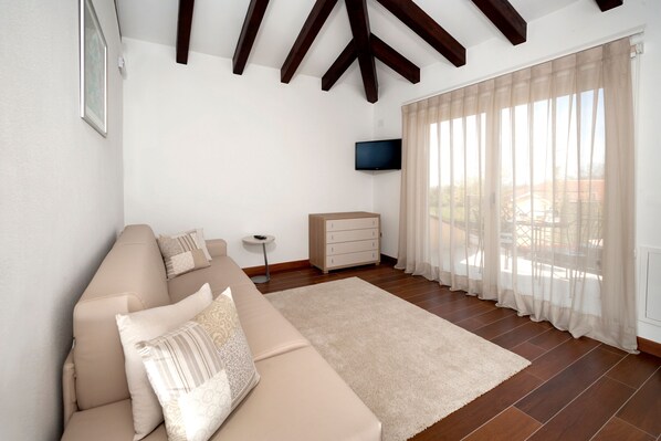 A living room with a beige sofa, a small white side table, a TV mounted on the wall, and a large window with sheer curtains.
