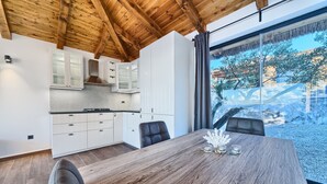 A modern kitchen with white cabinets and a wooden ceiling, a dining area with a wooden table and chairs, and a large window offering a view of trees and buildings. - Lux V in Murter With 2 Bedrooms and 1 Bathrooms (Tisno)