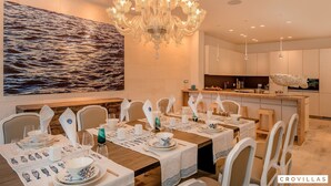 Un comedor con una gran araña, sillas blancas y una mesa larga dispuesta para una comida.