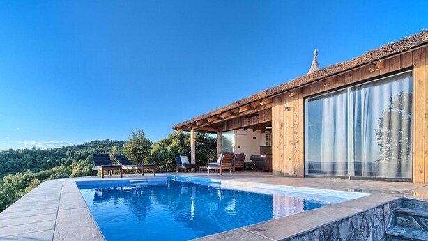 Uma área moderna de piscina ao ar livre com degraus de pedra que levam a um deck de madeira, cercada por vegetação exuberante e um céu azul límpido.