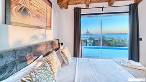 Quarto com uma janela grande, cama com almofadas estampadas, teto de madeira e vista para uma construção distante.