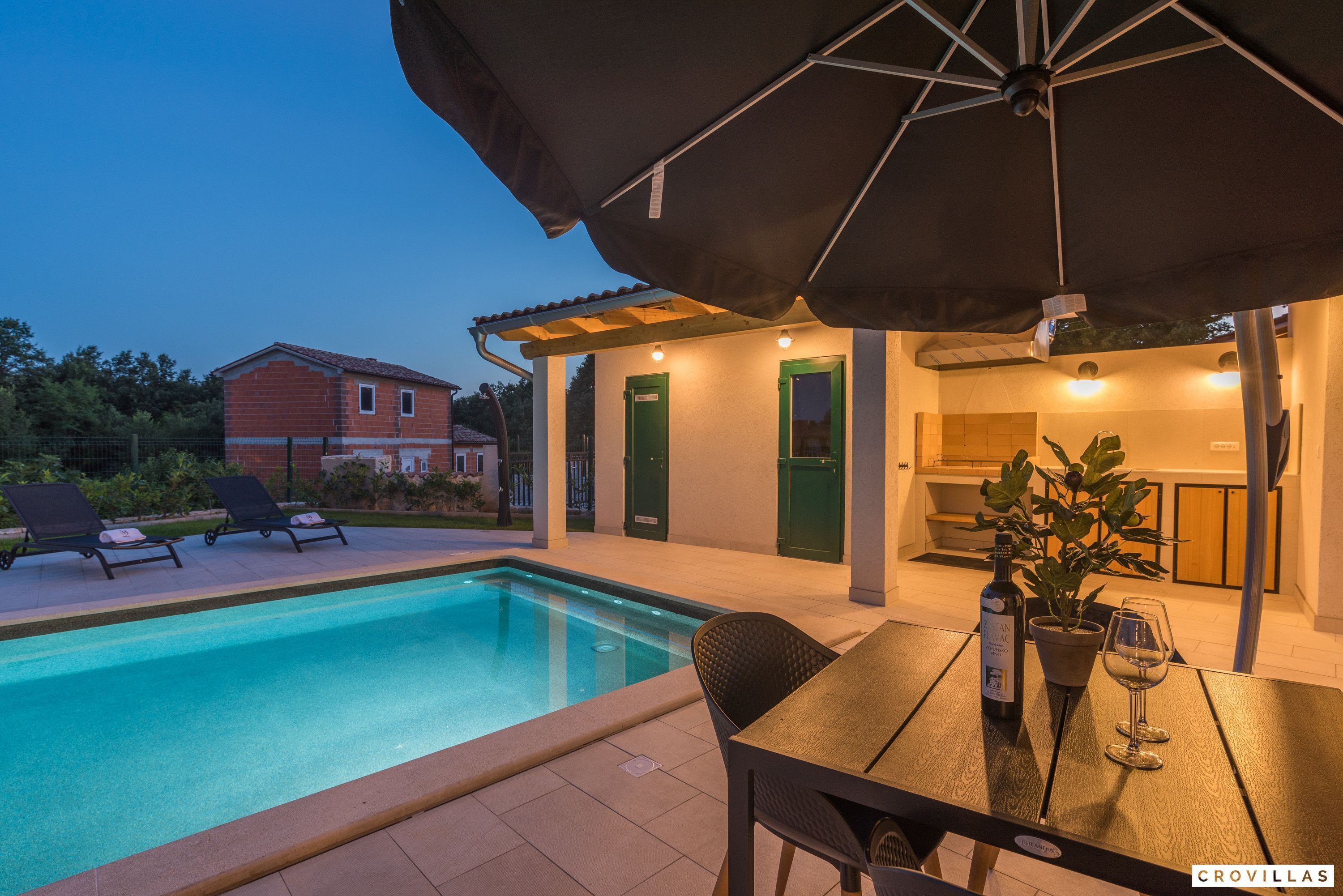 A poolside area with a table, chairs, and an umbrella. There are lounge chairs and a house in the background.