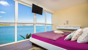 A bedroom with a large window overlooking a body of water, a bed with multiple pillows, a mounted TV, and a bedside table with a lamp. - Cama in Posedarje With 4 Bedrooms and 2 Bathrooms (Posedarje)