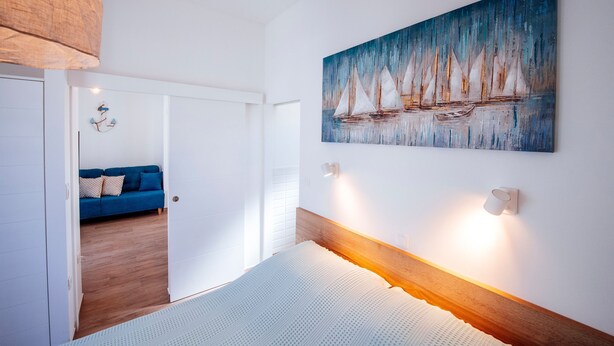 A bedroom with a bed, a painting of sailboats, and a blue sofa in the adjacent room. - Muntan in Kraj With 2 Bedrooms and 2 Bathrooms (Pasman)