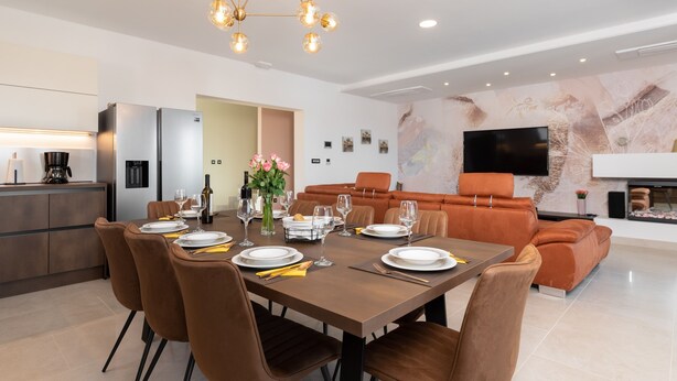 A modern dining area with a wooden table set for four, surrounded by brown upholstered chairs. A large, contemporary chandelier hangs above, and a flat-screen TV is mounted on the wall. - Callista in Ru i i (Sveta Nedelja)