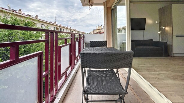 Un balcon avec une chaise en osier et une table, une porte vitrée donnant sur l’intérieur, et une vue sur les bâtiments en arrière-plan.