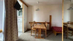 A room with a wooden dining table and chairs, a small bed with a red cover, a television mounted on the wall, and a ceiling light fixture. - 200m from resort center, 4th floor, view ski slopes, swimming pool, balcony, tv, ski locker, 28m² (La Plagne)