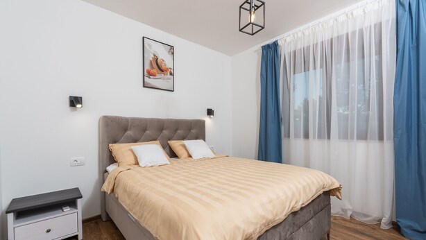 Un dormitorio con cama, mesitas de noche, lámpara de pared, un cuadro y una ventana con cortinas.