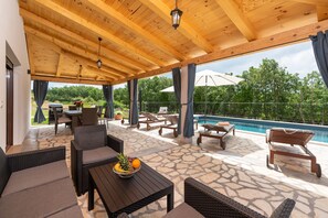 Eine Terrasse im Freien mit Korbmöbeln, Steinboden und einem Pool im Hintergrund.