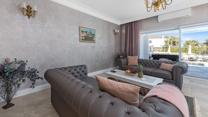 A modern living room with a grey sectional sofa, a grey armchair, and a coffee table. There is a large window with a view of a pool and outdoor furniture. - Carolus Dalmatia in Vodice (Vodice)