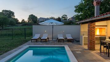 A swimming pool with lounge chairs and an umbrella, a brick fireplace, and a view of the surrounding area.