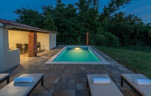 A rectangular swimming pool with a tiled deck, surrounded by lounge chairs with towels, a small covered seating area, and a brick pillar. - Matea Labin in Vadre (Barban)