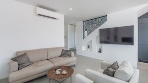 Una sala de estar moderna con un sofá color beige, una mesa de centro redonda y un sillón gris. Un televisor de pantalla plana está montado en la pared, y hay una escalera con un diseño geométrico.