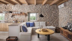 A rustic interior with a stone wall, wooden beams, a sofa with cushions, a round wooden table, and a kitchen area with a window. - Flavia in Umag With 4 Bedrooms and 3 Bathrooms (Umag)