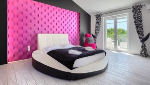 A modern bedroom with a curved bed, a pink accent wall, and a large window with black and white curtains.