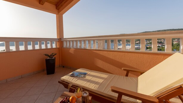A balcony with a wooden ceiling, a cushioned lounge chair, a small table with a magazine, a potted plant, and a view of a body of water and buildings in the distance. - Patria in Ra anj With 4 Bedrooms and 2 Bathrooms (Rogoznica)