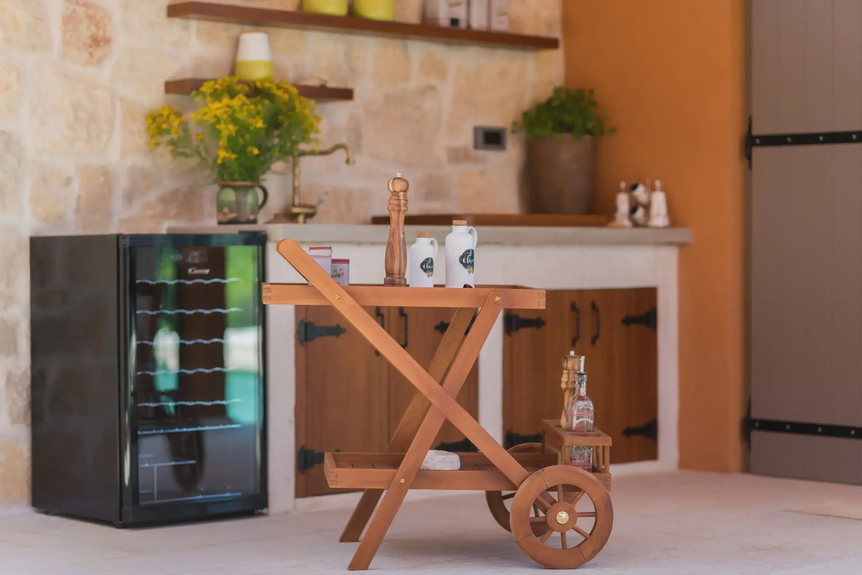 A kitchen with a wooden cart, a refrigerator, and a stone wall.