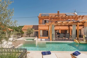 A pool area with lounge chairs, a dining area, and a building with a tiled roof and solar panels.