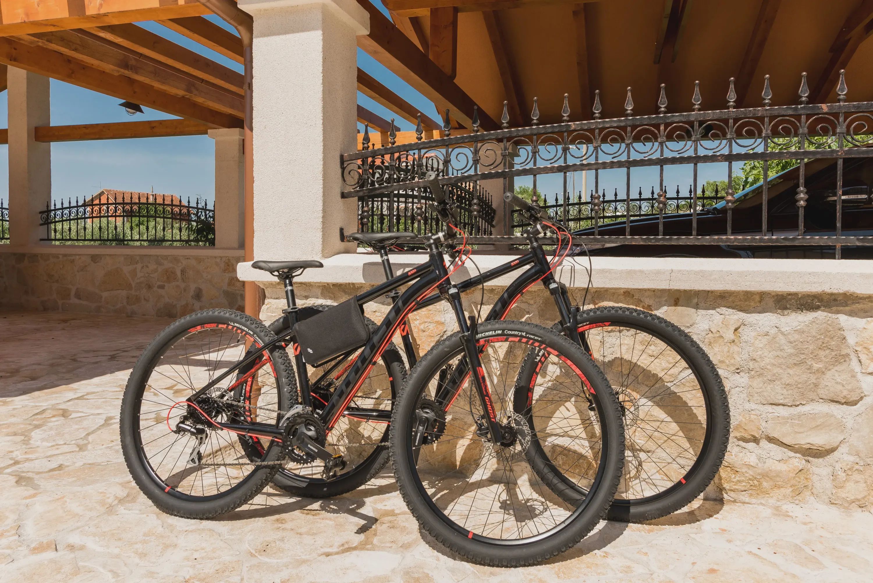 Un vélo est garé sous une pergola en bois, avec des murs en pierre et une clôture en fer forgé en arrière-plan.