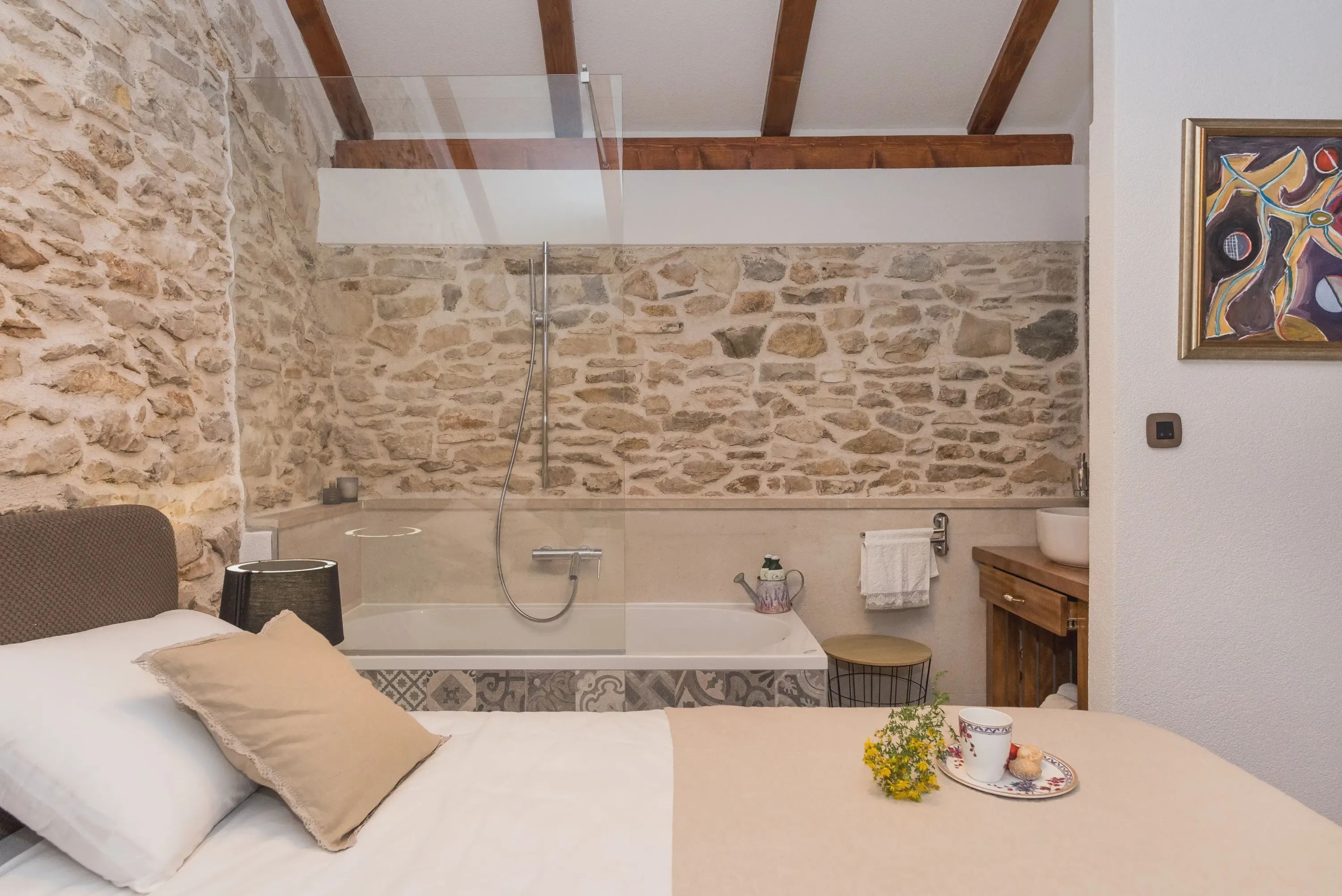 Un dormitorio con pared de piedra, una cama con almohadas, cabina de ducha de vidrio, bañera, una pequeña mesa con una taza y flores, y un armario de madera.
