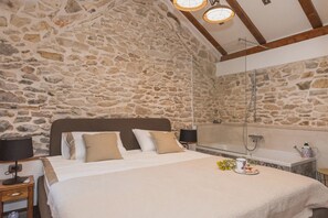Ein Schlafzimmer mit Steinwand, Holzboden, einem Bett mit weißer Bettwäsche, zwei Nachttischen mit Lampen sowie ein Badezimmer mit Badewanne und Dusche.