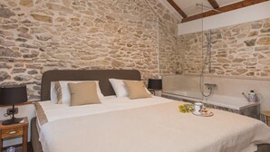 Une chambre avec un mur en pierre, un plafond en bois, un lit avec une literie blanche, deux tables de chevet avec des lampes, et une salle de bain avec une baignoire et une douche.