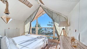 A cozy bedroom with a wooden floor, a bed with white linens, a wooden table with a lamp, and a large window offering a view of trees and a lake. - Treehouse in Murter With 1 Bedrooms and 1 Bathrooms (Tisno)