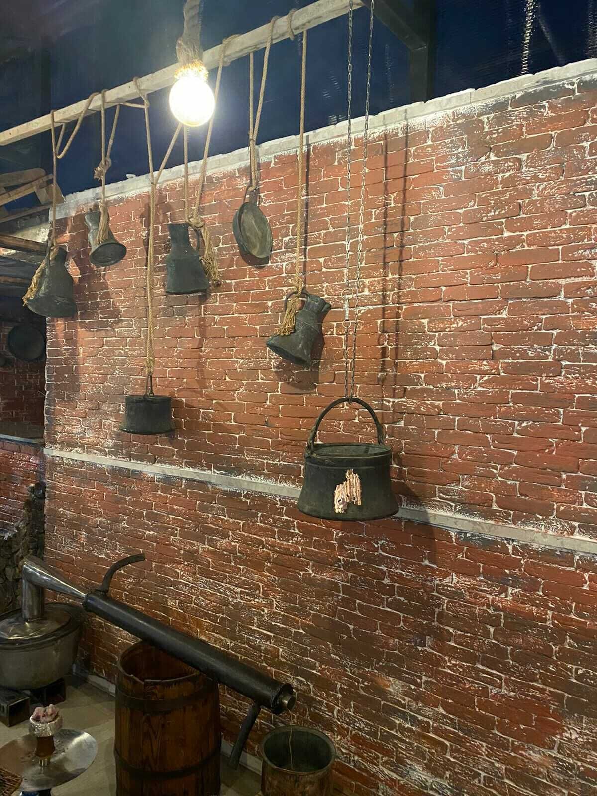 A rustic interior with exposed brick walls, hanging metal pots, and a large metal barrel.