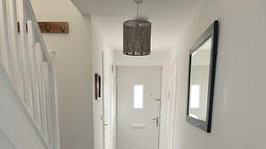 A hallway with a wooden floor, a grey rug, a white bench, a staircase, a door with a window, a wall-mounted mirror, and a ceiling light fixture.