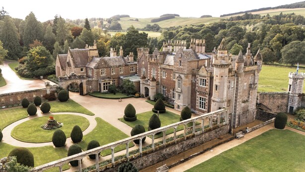 A large, historic castle with multiple towers, a well-maintained garden, and a bridge leading to the entrance.