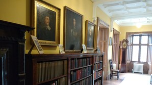 A library with bookshelves, framed portraits, a fireplace, and a large window.