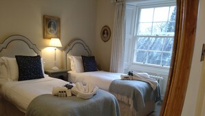 A hotel room with two beds, each with a white headboard, a bedside table with a lamp, and a window with a view of the outside.