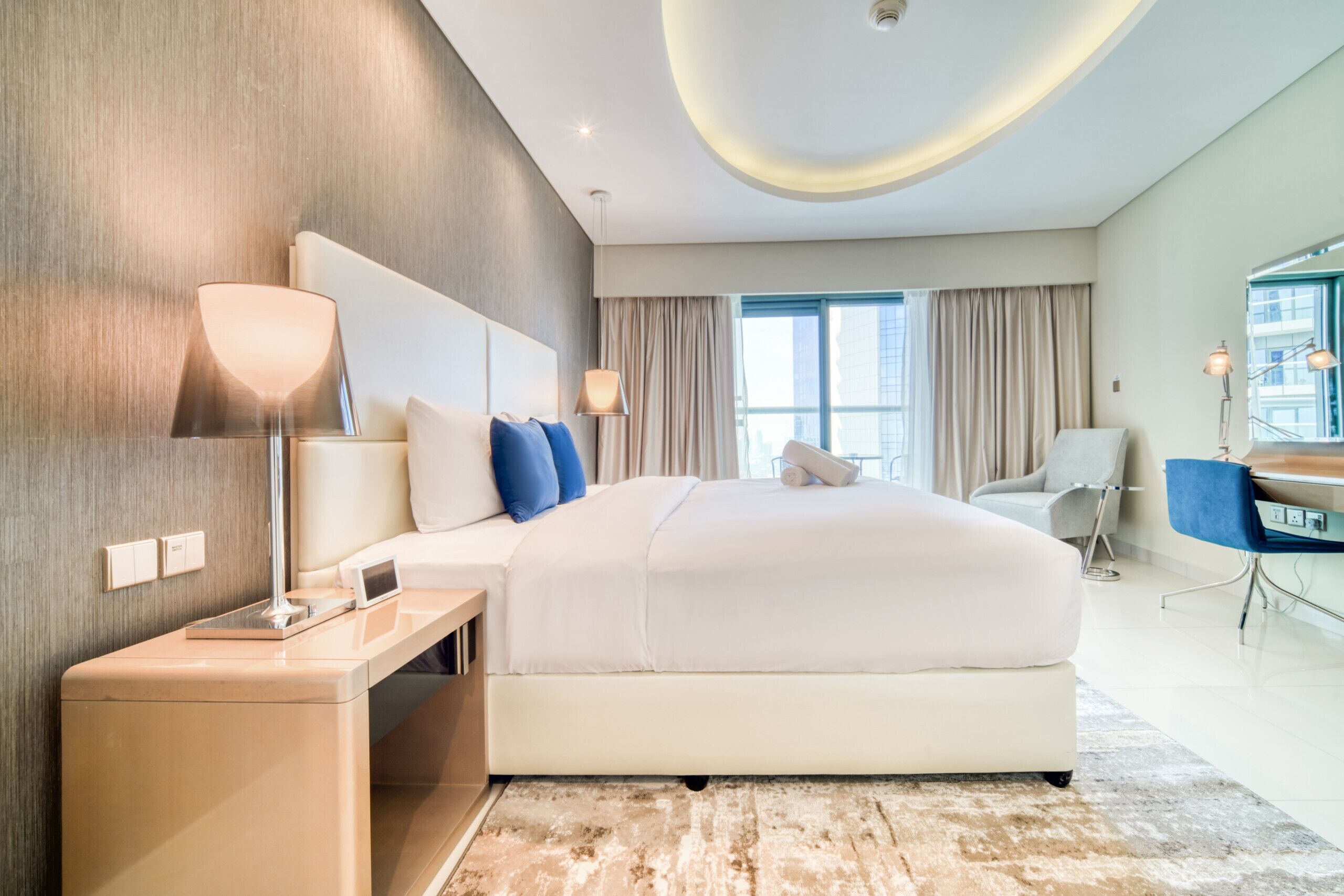Una camera d'albergo moderna con un letto grande, lampade ai comodini, una scrivania e vista sulla città attraverso ampie finestre.