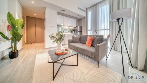 A modern living room with a grey sofa, a coffee table, and a floor lamp. There is a kitchen area in the background with white cabinets and a dining table. - Picturesque Studio at Mesk 1 Midtown Dubai Production City by Deluxe Holiday Homes (Dubai)