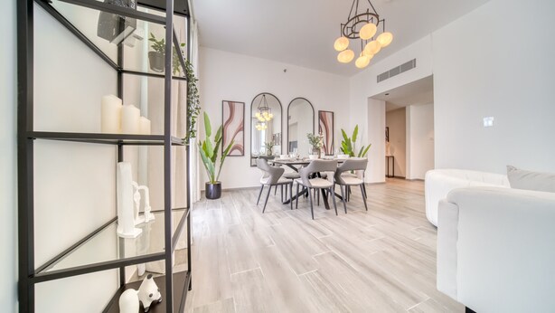 A modern living room with a white sofa, a dining area with a table and chairs, and a staircase with a black railing. - Harmonious 2BR w/ Assistant’s Room at Mesk 1 Midtown Dubai Production City by Deluxe Holiday Homes (Dubai)