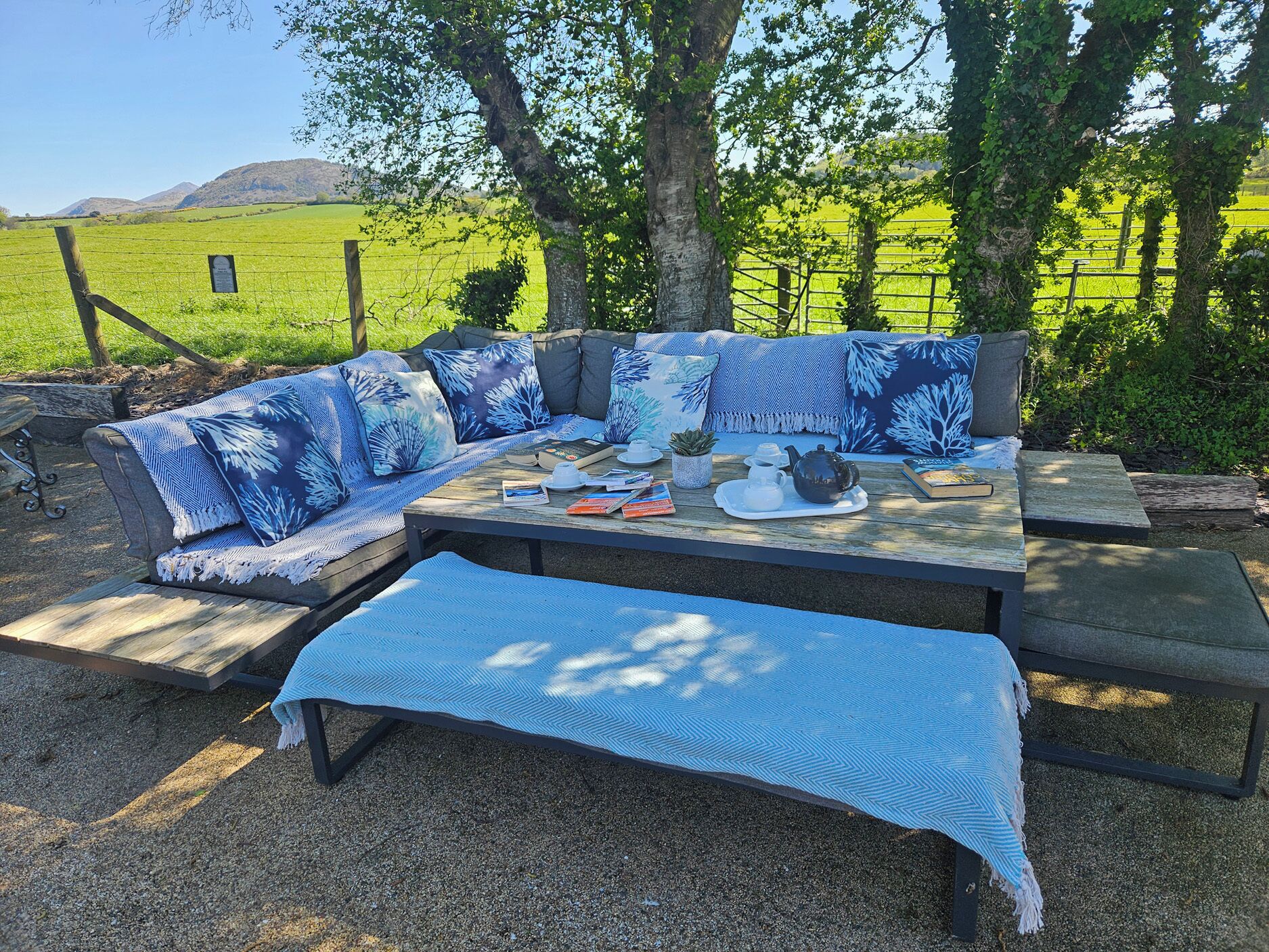 Un espacio al aire libre con sofá y banco, una mesa con tetera y libros, y vistas a un campo verde y colinas.