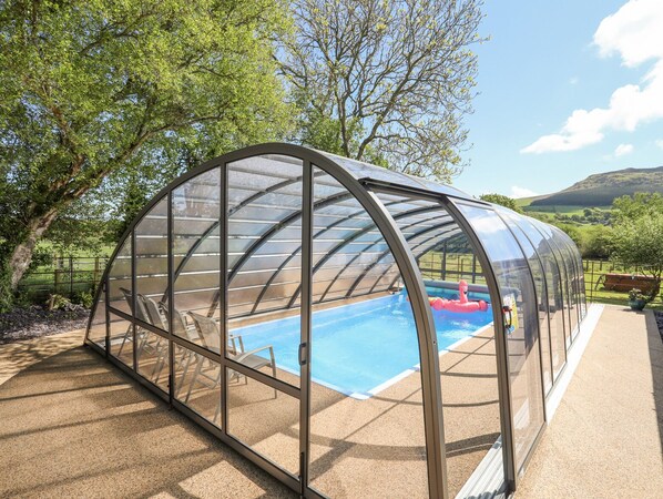 A glass-enclosed swimming pool with a curved roof, surrounded by trees and a view of hills.