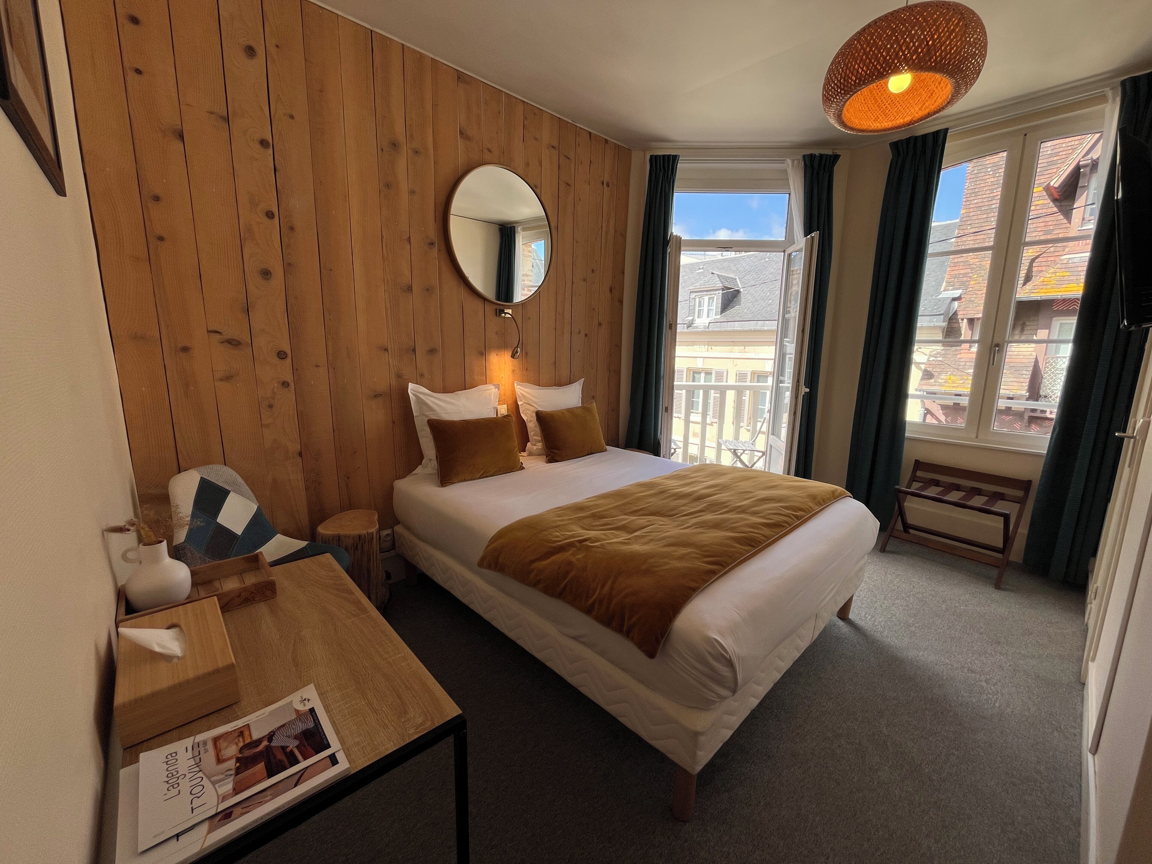 A hotel room with a wooden wall, a bed with white and brown bedding, a bedside table with a lamp, a window with curtains, and a view of buildings outside.