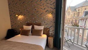 Een slaapkamer met een bed, kussens, een houten hoofdbord, een nachtkastje, een rond houten krukje, en een balkon met een hekwerk.