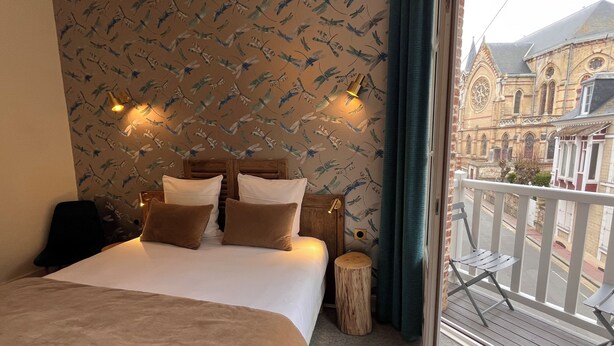 Une chambre à coucher comprenant un lit, des oreillers, une tête de lit en bois, une table de chevet, un tabouret rond en bois et un balcon avec une balustrade.