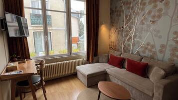 A living room with a sofa, a coffee table, a TV on a stand, a radiator, a window with curtains, and a wall with a tree pattern.